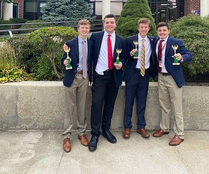 Four students in blazers holding trophies outside a tournament venue